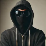 A person wearing a black hooded jacket, black sunglasses, and a black balaclava against a neutral background.